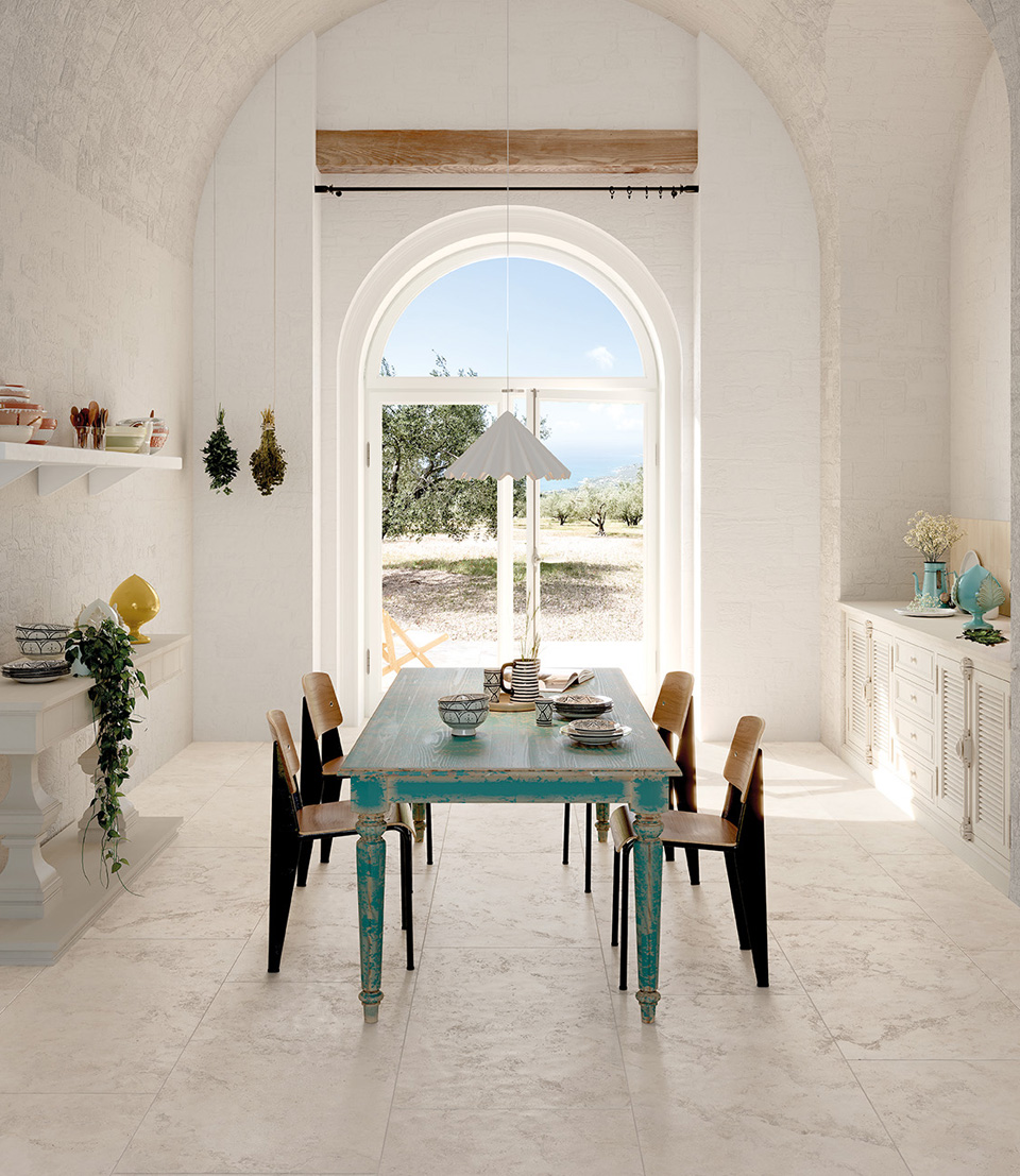 Cucina dal gusto mediterraneo con pavimento in gres chiaro, pareti bianche, tavolo azzurro decapato e ampie finestre ad arco con vista esterna.