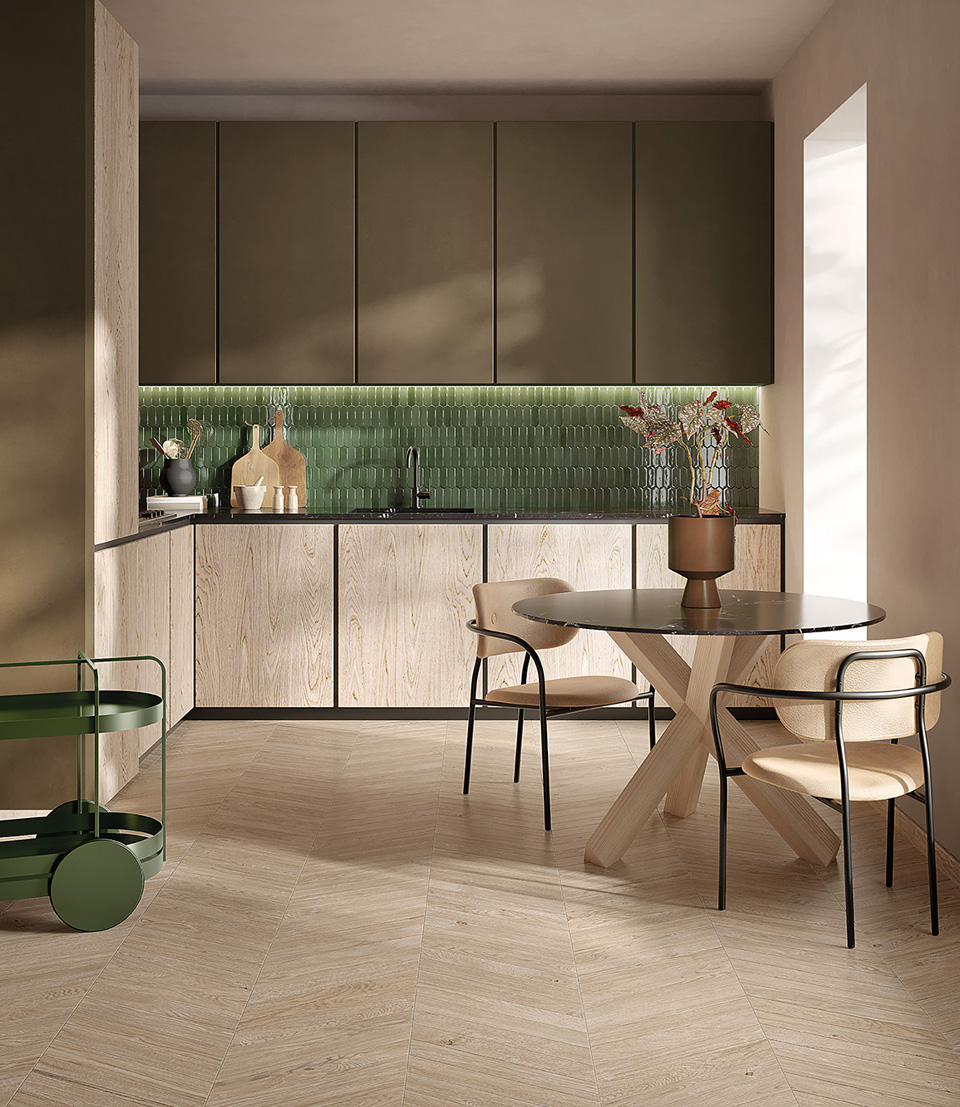 Cucina moderna con backsplash verde lucido, arredi in legno chiaro, tavolo tondo con base a croce e sedute imbottite beige.